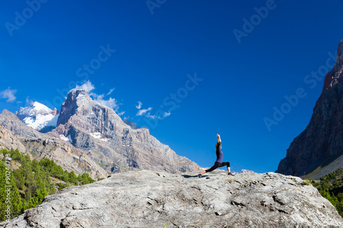Young woman doing exercise Cute Girl Doing Yoga Fitness to Stretch Body Staying on High Rock at Mountain Panoramic Landscape Outdoor Sunny Sky Peaks Wilderness Country Smiling Beautiful Face