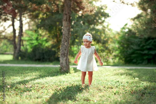 Beautiful baby girl walking in park. Wearing trendy white dress outdoors. Childhood. photo