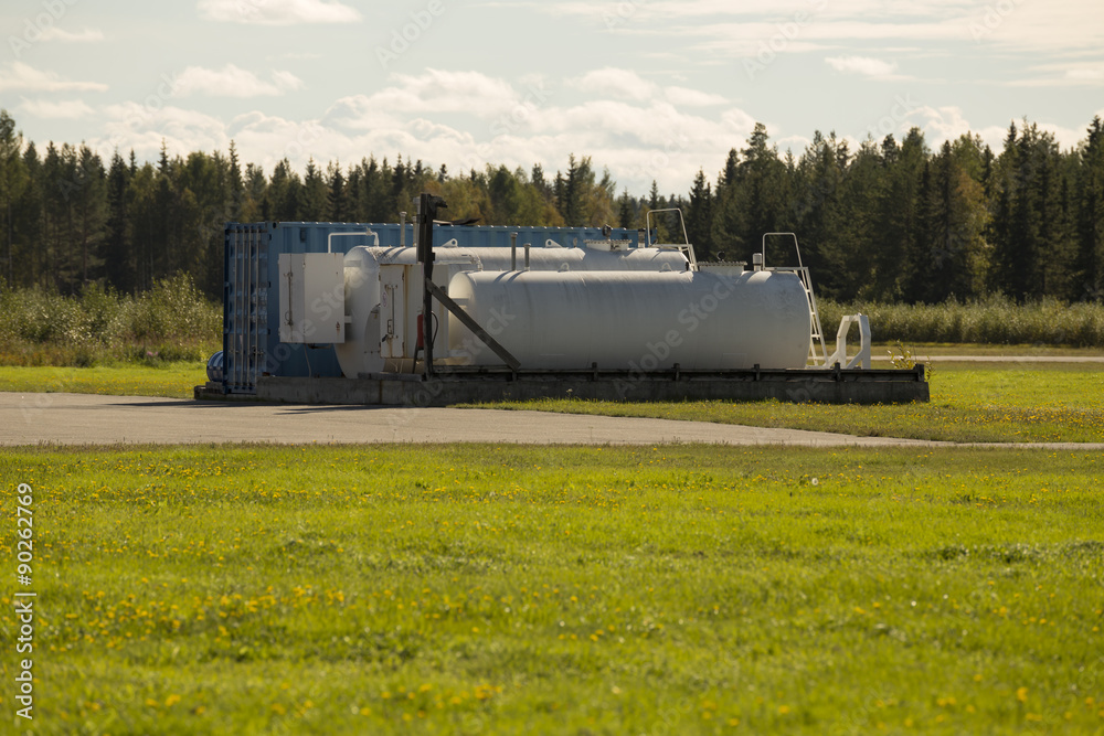 Fuel tanks on an air field with some grass in the foreground and forest in the background