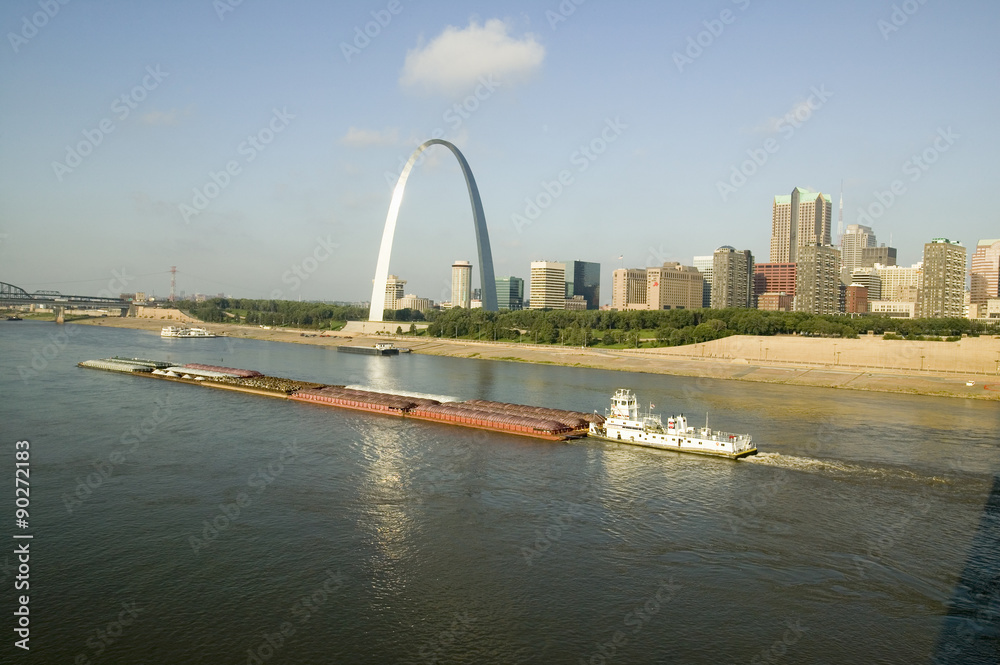 Obraz premium Daytime view of tug boat pushing barge down Mississippi River in front of Gateway Arch and skyline of St. Louis, Missouri as seen from East St. Louis, Illinois on the Mississippi River