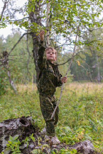 The boy in the autumn forest camouflage with a stick in his hand on an old tree stump