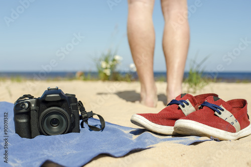 Camera with lens lying a towel on the sand at the beach, standing beside red shoes, and in the background can be seen the feet of the photographer, looking at the sea
