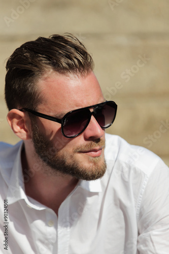Portrait of a good looking man in his 20s, wearing a white shirt with dark sunglasses, sitting down outside on a set of steps on a sunny summer day.