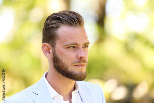 Portrait of a man in his 20s wearing a light blue blazer and white shirt, standing against a green background on sunny summer day. 