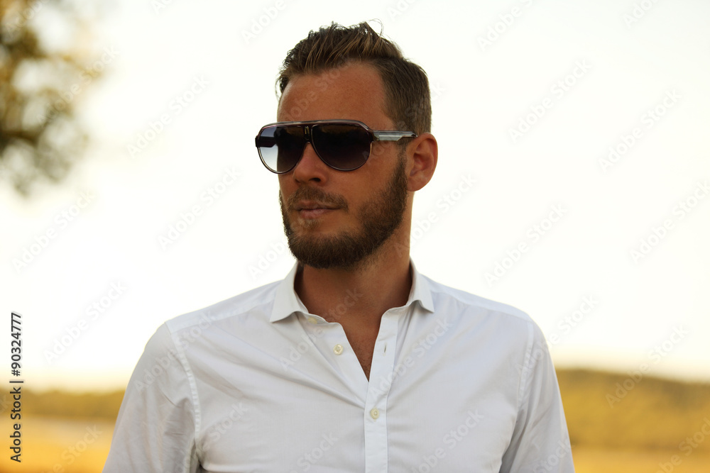 A man wearing a white shirt and dark sunglasses, standing against a large yellow field looking away from camera. A great sunny summer day. A man wearing a white shirt and dark sunglasses, standing against a large yellow field looking away from camera. A great sunny summer day.