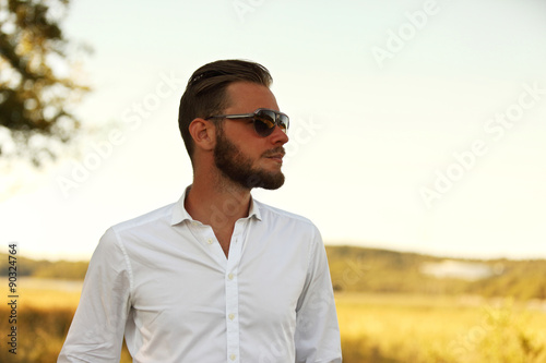 A man wearing a white shirt and dark sunglasses, standing against a large yellow field looking away from camera. A great sunny summer day.