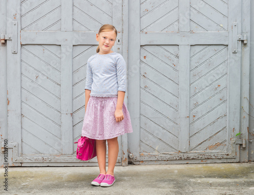 Fashion portrait of a cute little girl of 8 years old, wearing grey sweatshirt, pink skirt and laceless shoes, holding a bag, outdoors photo