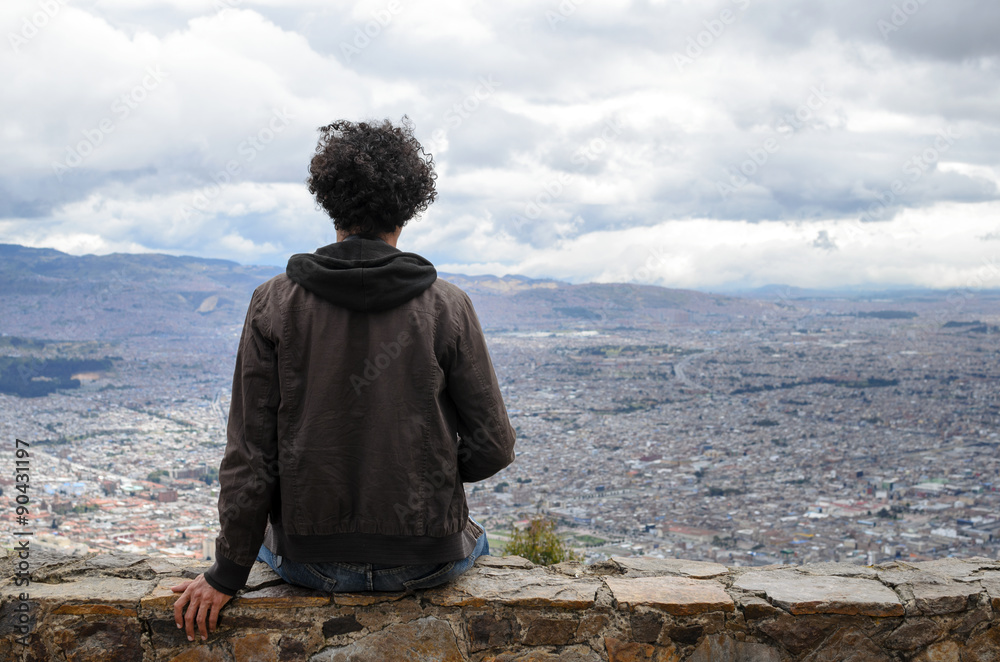 Anonymous guy with black curly hair sitting on stone wall overlooking Bogota city 