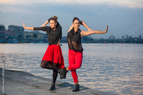 Two young beautiful twin sisters are dancing waacking dance in the city background near river. showing the different style and pose of modern dance with black and red dress near water on summer time. photo