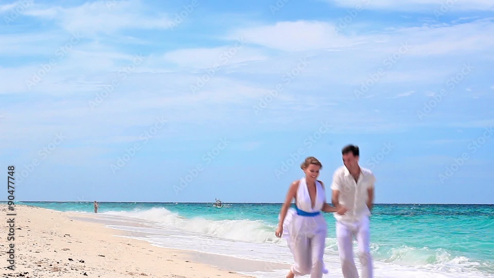 Young couple holding hands running on tropical sandy beach in white dresses
Young couple holding hands running on tropical sandy beach in white dresses