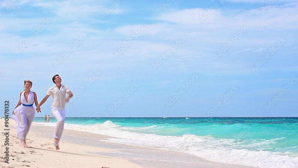 Young couple holding hands running on tropical sandy beach in white dresses
Young couple holding hands running on tropical sandy beach in white dresses