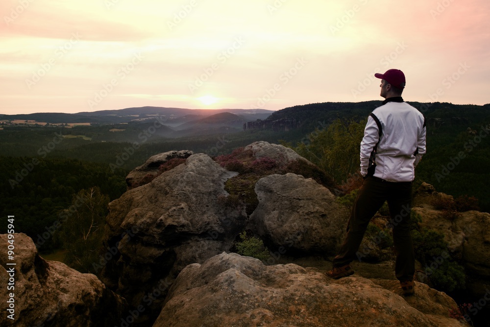 Sportsman with red baseball cap and in white jacket on rocky peak. Dreamy fogy landscape, beautiful valley below Sportsman with red baseball cap and in white jacket on rocky peak. Dreamy fogy landscape, beautiful valley below