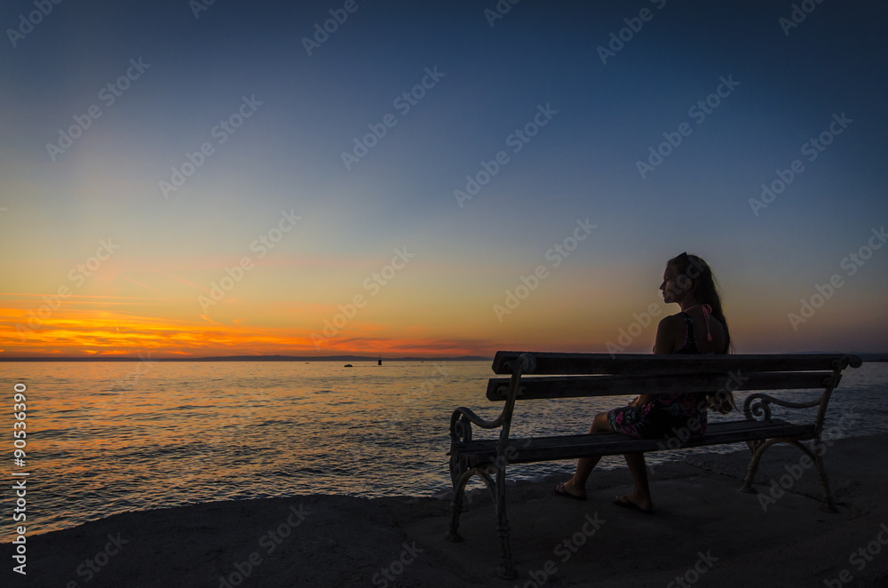 Young girl sitting on bench and looking at sunset over the sea