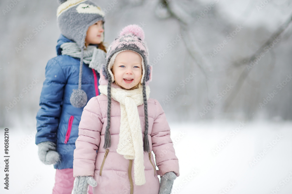 Two funny adorable little sisters having fun together in beautiful winter park during snowfall