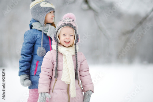 Two funny adorable little sisters having fun together in beautiful winter park during snowfall