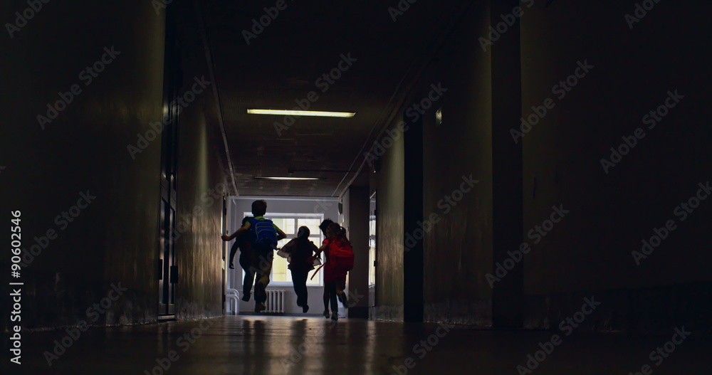Rear view of elementary students running along dark school corridor 
