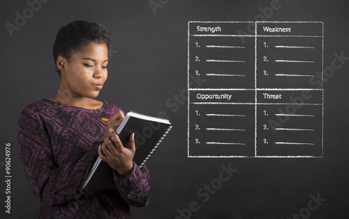African American woman writing in book diary SWOT analysis on blackboard background