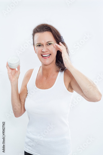 Aufgeregte Brünette Frau trägt weiße Muskelshirt, hält ein Kosmetikglas und wendet Feuchtigkeitscreme im Augenbereich an, Studioaufnahme mit weißem Hintergrund
