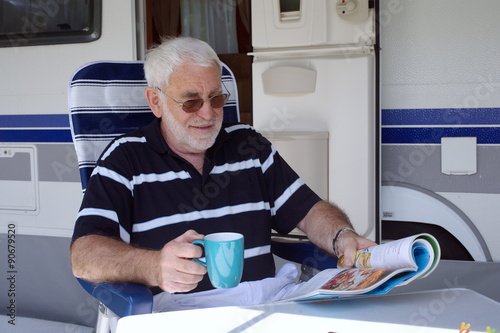 Rentner sitzt Kaffee trinkend  vor seinem Wohnmobil oder Wohnwagen und liest photo