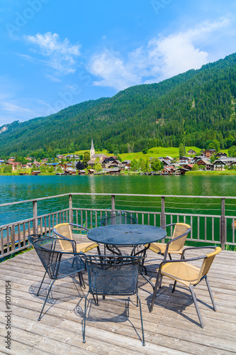 Table with chairs on wooden deck and view of green water Weissensee lake in summer landscape of Alps mountains, Austria