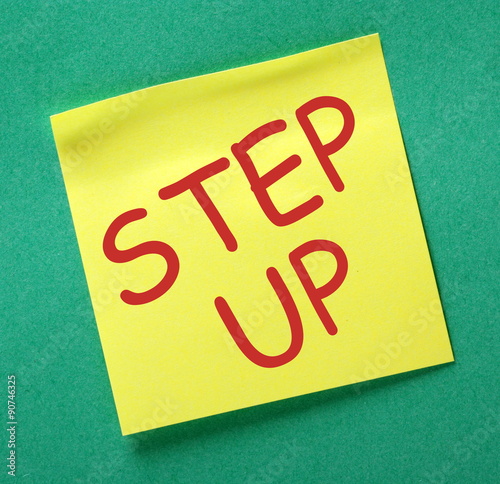 The phrase Step Up in red text on a yellow sticky note posted on a green notice board as a reminder to make a contribution photo