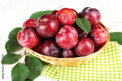 Ripe plums in wicker bowl on wooden table with napkin, closeup