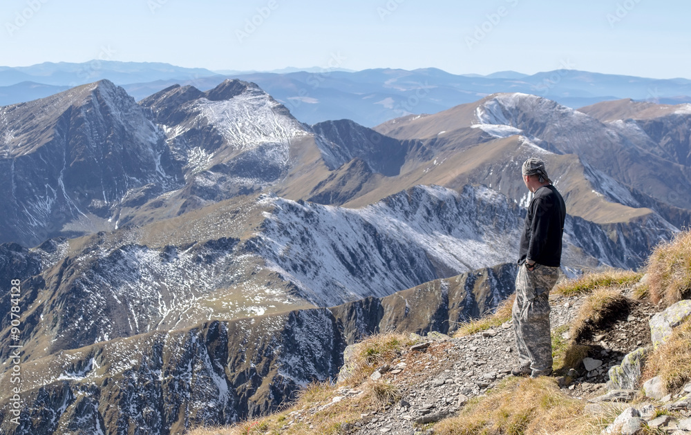 Lonely man stands on top of a mountain and looking into the distance on a background of mountain range. Focus on the first plan, on the man. Mountains on the background is blurry.
