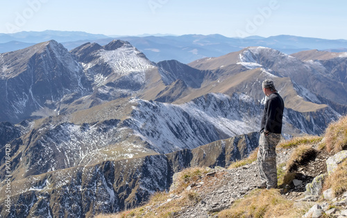 Lonely man stands on top of a mountain and looking into the distance on a background of mountain range. Focus on the first plan, on the man. Mountains on the background is blurry. © oleksandrmazur