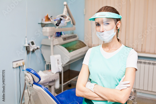 Young attractive woman dentist in protective workwear with protection face shield standing in front of dentist chair with hands crossed with confidence