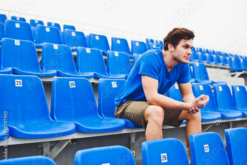 Man athlete in blue shirt sits relaxing alone on chairs on stadium after hard workout. Massaging injured hand after training