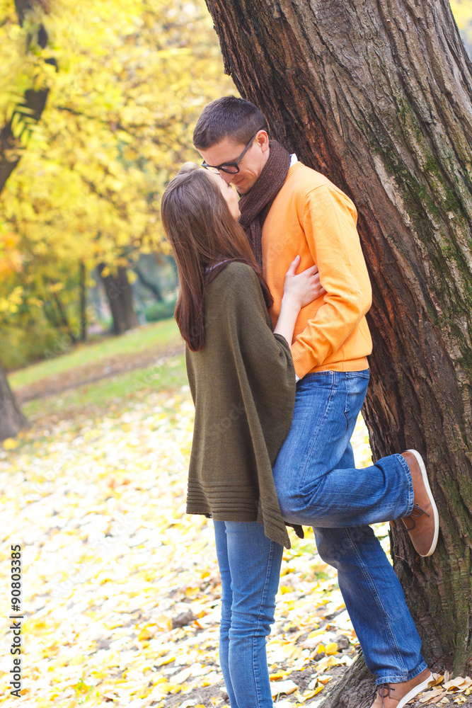 Fototapeta premium Young man kissing a girl in a park on a sunny autumn day while leaning on a tree.