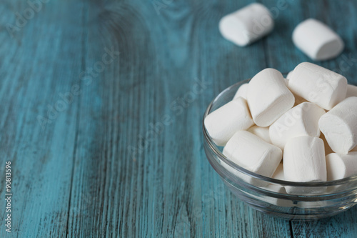 White soft marshmallows in glass bowl on blue wooden background, selective focus