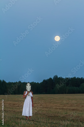 Scaring young woman wearing white dress with horse skull on her head at night field