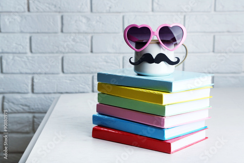 Books and cup with mustache on wooden table on brick wall background