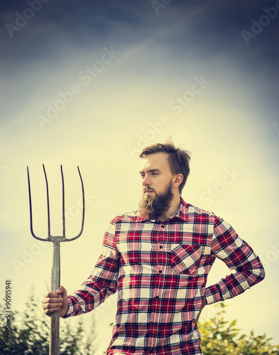 Portrait of young bearded farmer in red checkered shirt with old pitchfork on sky  nature backgrund, toned