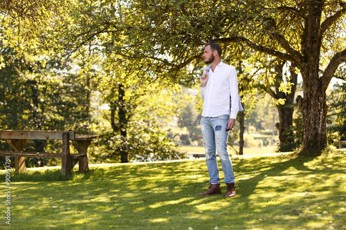 A man standing on green grass with trees behind him, wearing a white shirt and jeans, and holding a blazer on his shoulder. A sunny summer day.