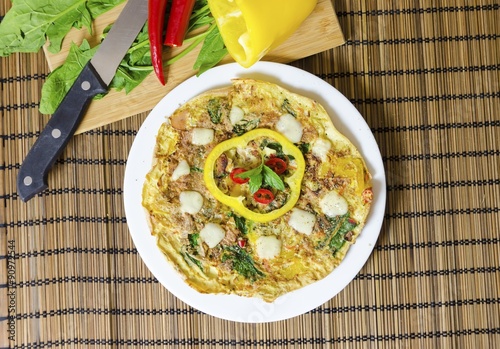 A spinach, chili, mint and pepper omelette in a dish over bamboo table cloth and chopping board with knife and raw vegetables on while background. A healthy diet meal omelet. photo