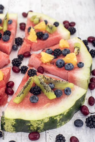 colorful tropical fruit watermelon pizza topped with kiwifruit, blueberries, orange and fresh berries cut into segments on a rustic wooden board