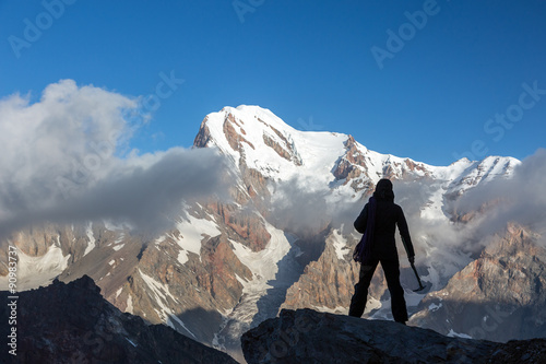 Alpine Climber Reached Summit Silhouette Woman Staying on Top of Rock Cliff Holding Climbing Gear Stormy Clouds and Peaks Illuminated bright Morning Sun © alexbrylovhk