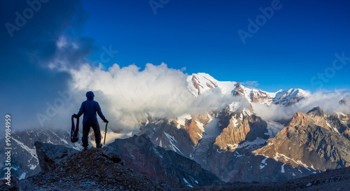 Alpine Climber Reached Summit Silhouette Man Staying on Top of Rock Cliff Holding Climbing Gear Stormy Clouds and Peaks Illuminated bright Morning Sun
