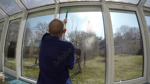 worker man with sun glasses spray liquid cleaner on glass and wipe window with washer squeegee tool in front of sun. Cleaning greenhouse windows in spring. 4K UHD wide angle shot.
 photo