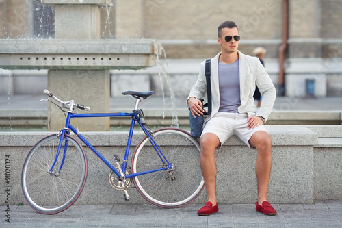 Handsome young man sitting near the fountain with his bicycle beside him