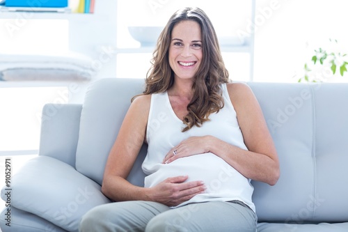 Smiling pregnant woman sitting with hand on stomach on sofa Free Download Smiling pregnant woman sitting with hand on stomach on sofa - GFXLoad