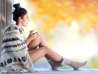Beautiful young smiling brunette woman with cup of coffee wearing knitted nordic print poncho sitting home by the window.. Blurred fall garden background. 