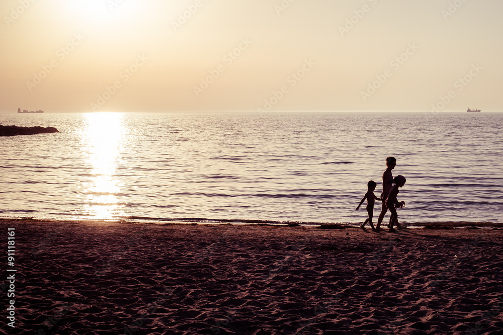 A silhouette of a mother and two children walking on a sand beach during the sunset in Albania on the Adriatic sea coast. The sea is calm with the sunlight reflection and the sky is cloudless.