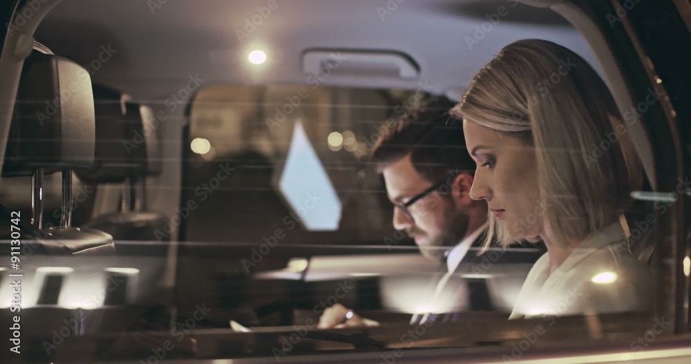 Businesswoman making phone call while her colleague using tablet computer in back seat of moving car 