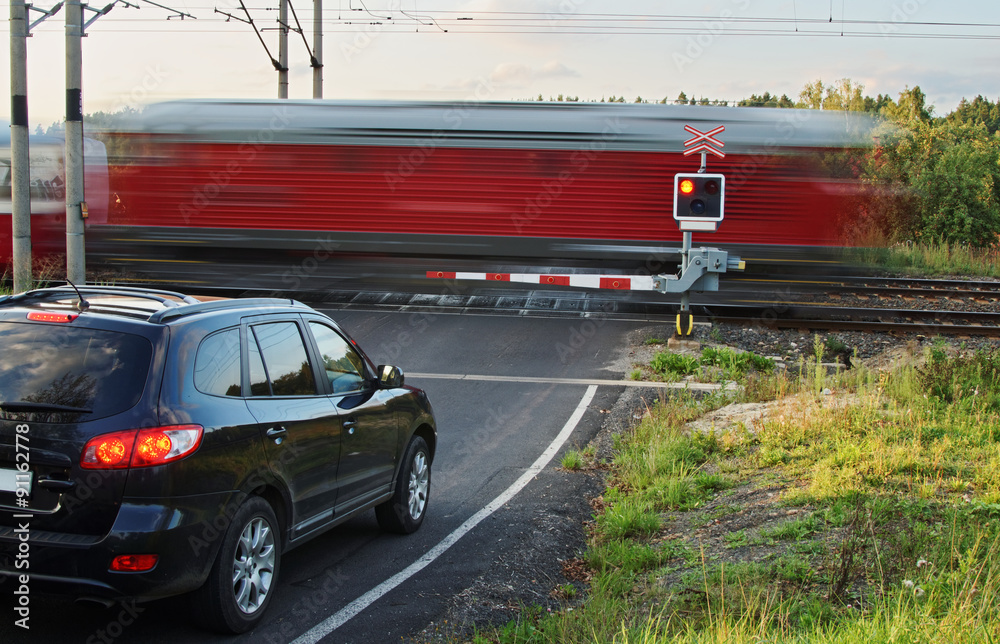 Obraz premium Speeding motion blur red train passing through a railway crossing with gates. Black car standing in front of the railway barriers on an asphalt road.