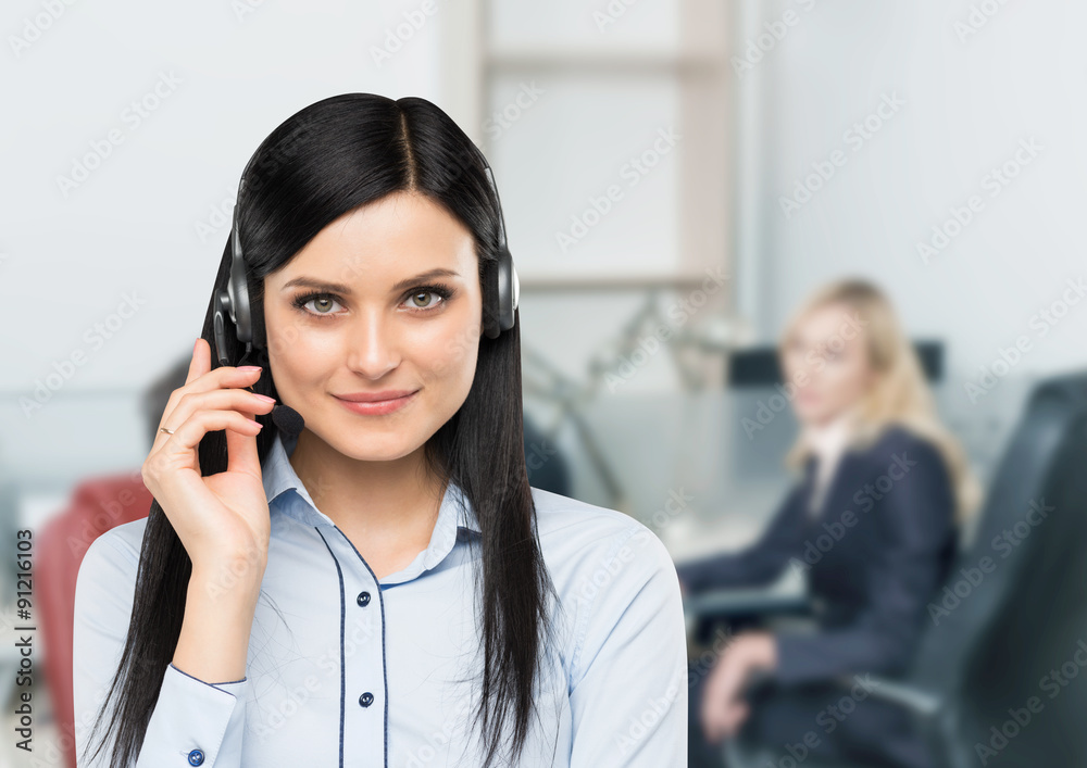 Front view of the smiling brunette support phone operator with headset. Office workplace background in blur.