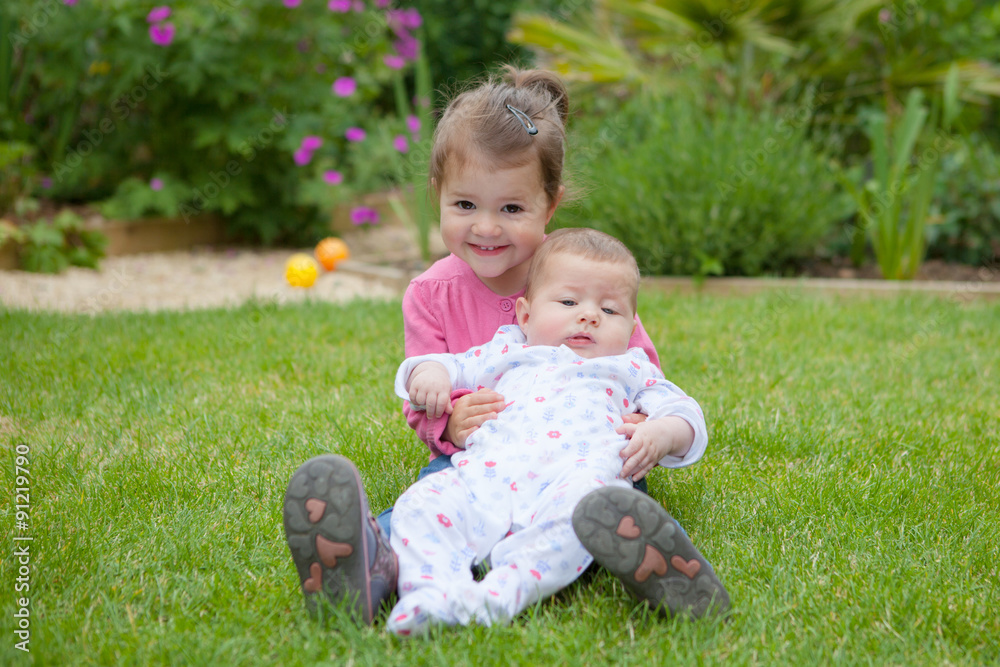 A little girl in the garden sitting on the grass in the garden hugging her baby sister A little girl in the garden sitting on the grass in the garden hugging her baby sister