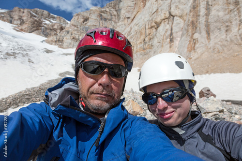 Joyful Alpine Climbers Self Portrait  Faces of Smiling Man and Woman Sport Style Clothing Protection Helmets Sunglasses High Altitude Mountain Landscape with Rock and Snow on Background © alexbrylovhk